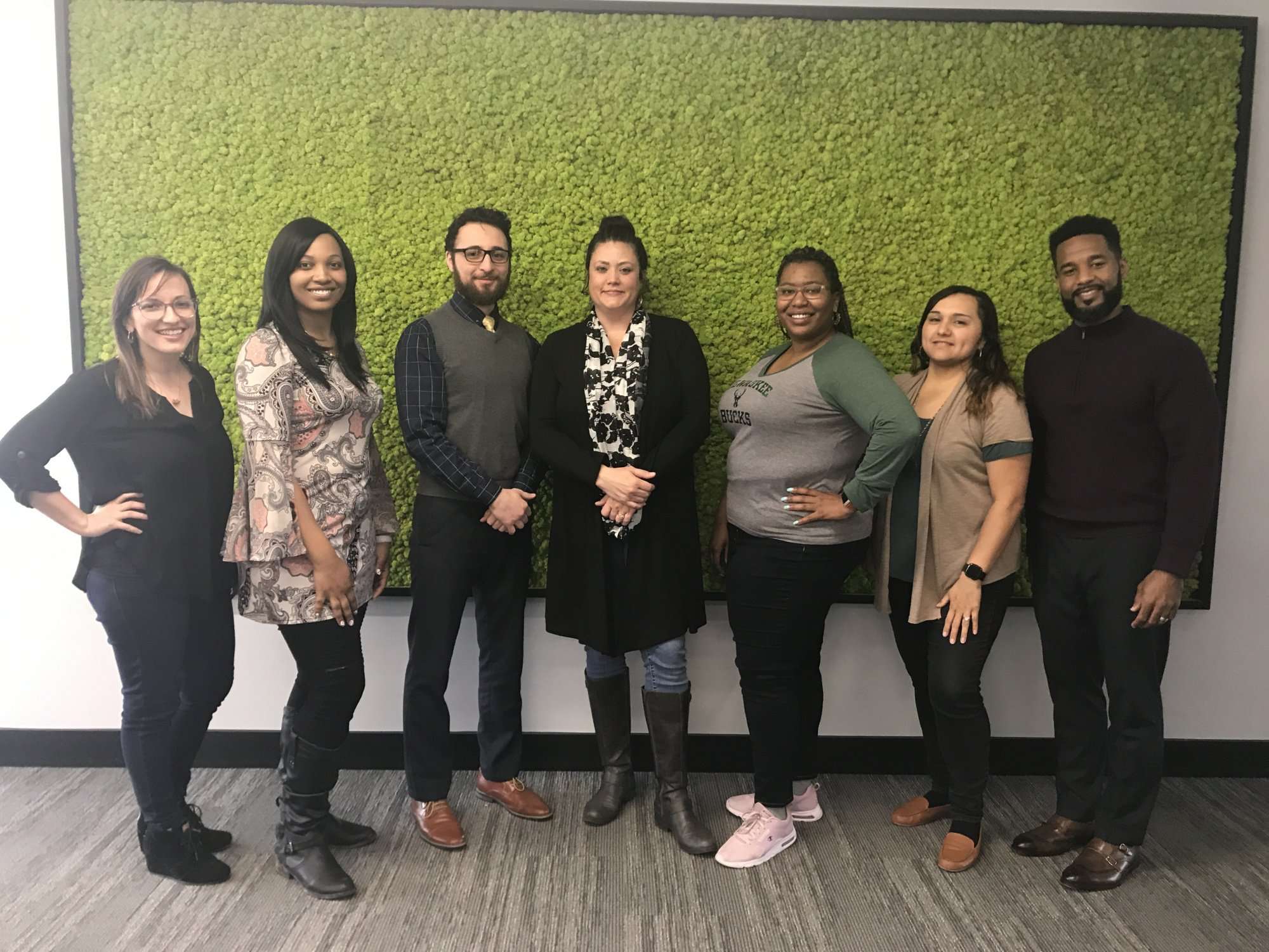 A group of seven people stand side by side, smiling in front of a green, moss-covered wall in an office environment.