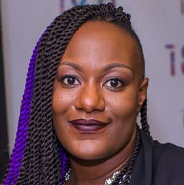 A person smiles while facing the camera, sporting purple and black braided hair. They wear dark lipstick and a dark top. The background includes a partially visible text banner.