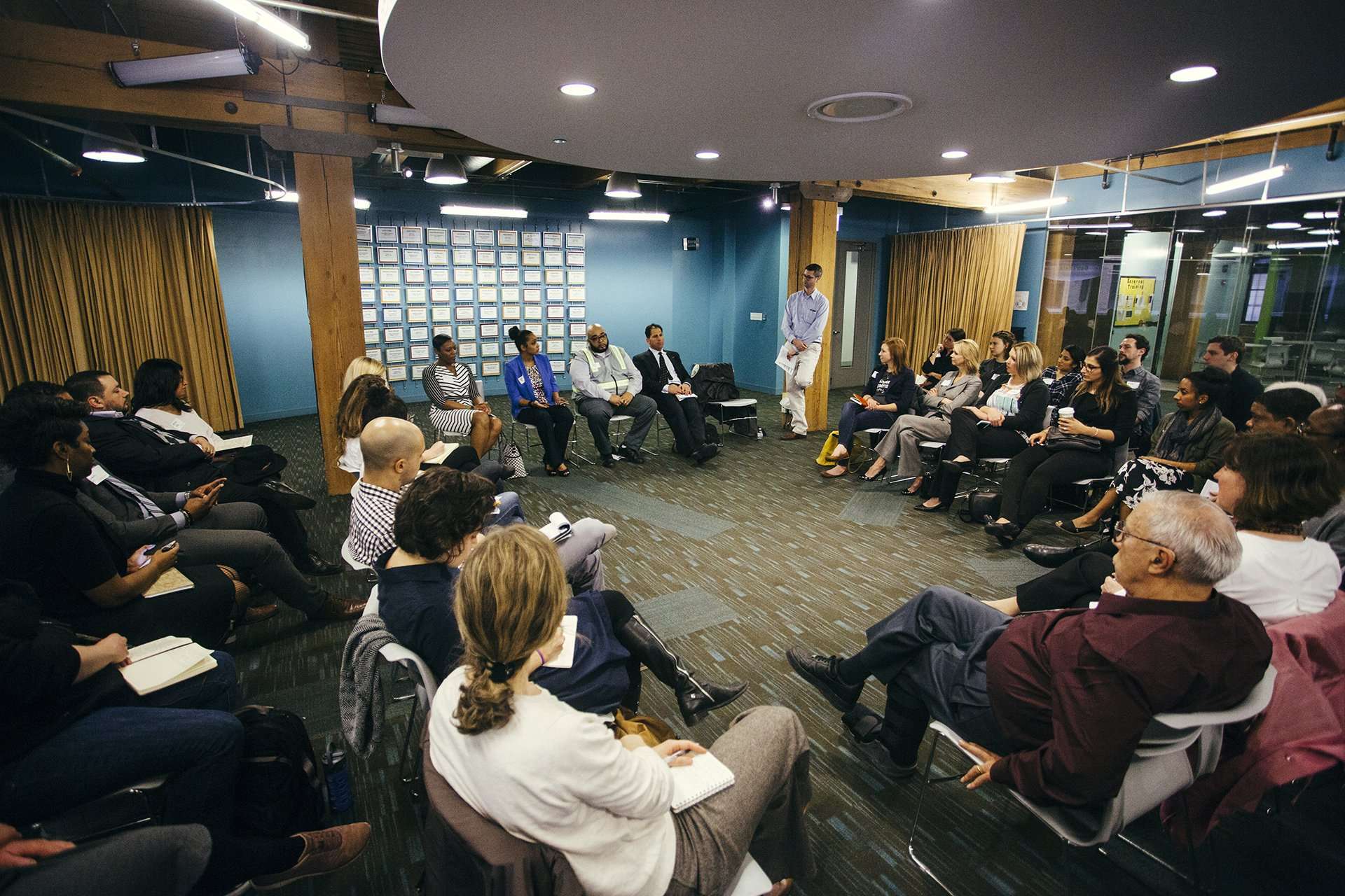 A diverse group of people seated in a circular arrangement participate in a discussion in a modern, lighted conference room with wood columns, glass walls, and a notice board in the background.