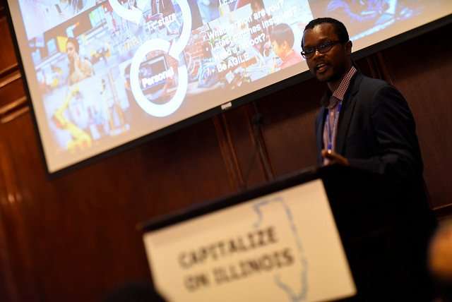 A man is speaking at a podium in a conference room, with a presentation screen behind him. The podium sign reads "CAPITALIZE ON ILLINOIS" and features an outline of the state.