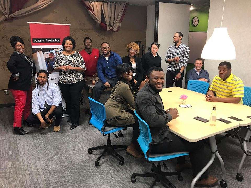 A group of 13 people, some standing and some seated around a table, are smiling in an office space with blue chairs. A banner reads, "i.c.stars // columbus Transforming Professionals Talent Technology Values-Based LEADERSHIP."