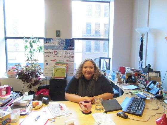A person smiles while seated at a cluttered office desk, holding a mug. Surrounding items include a computer, papers, and a poster listing companies like Accenture and USB, with large windows in the background.