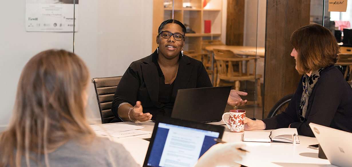 A woman is speaking while gesturing with her hands, seated at a round table with a laptop, surrounded by colleagues in a modern office with wooden furnishings.