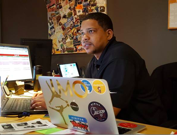 A man works at a desk using a laptop adorned with multiple stickers in a cluttered office, surrounded by papers, a coffee cup, and bulletin boards covered with images.