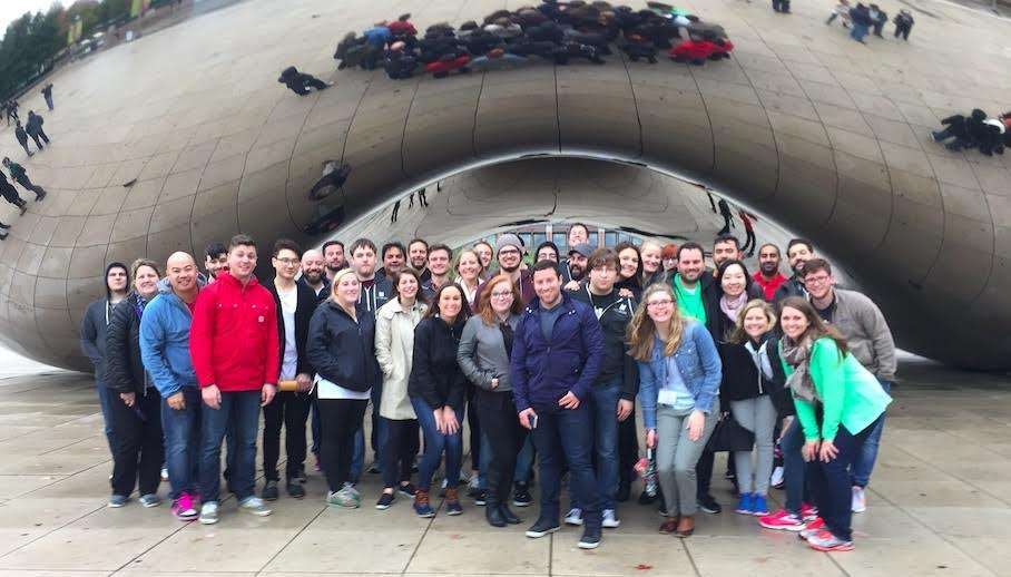 A group of around 30 people stands closely together, smiling, beneath the reflective, metallic sculpture "Cloud Gate" (The Bean) in Chicago's Millennium Park. Reflected images can be seen on the sculpture’s surface.