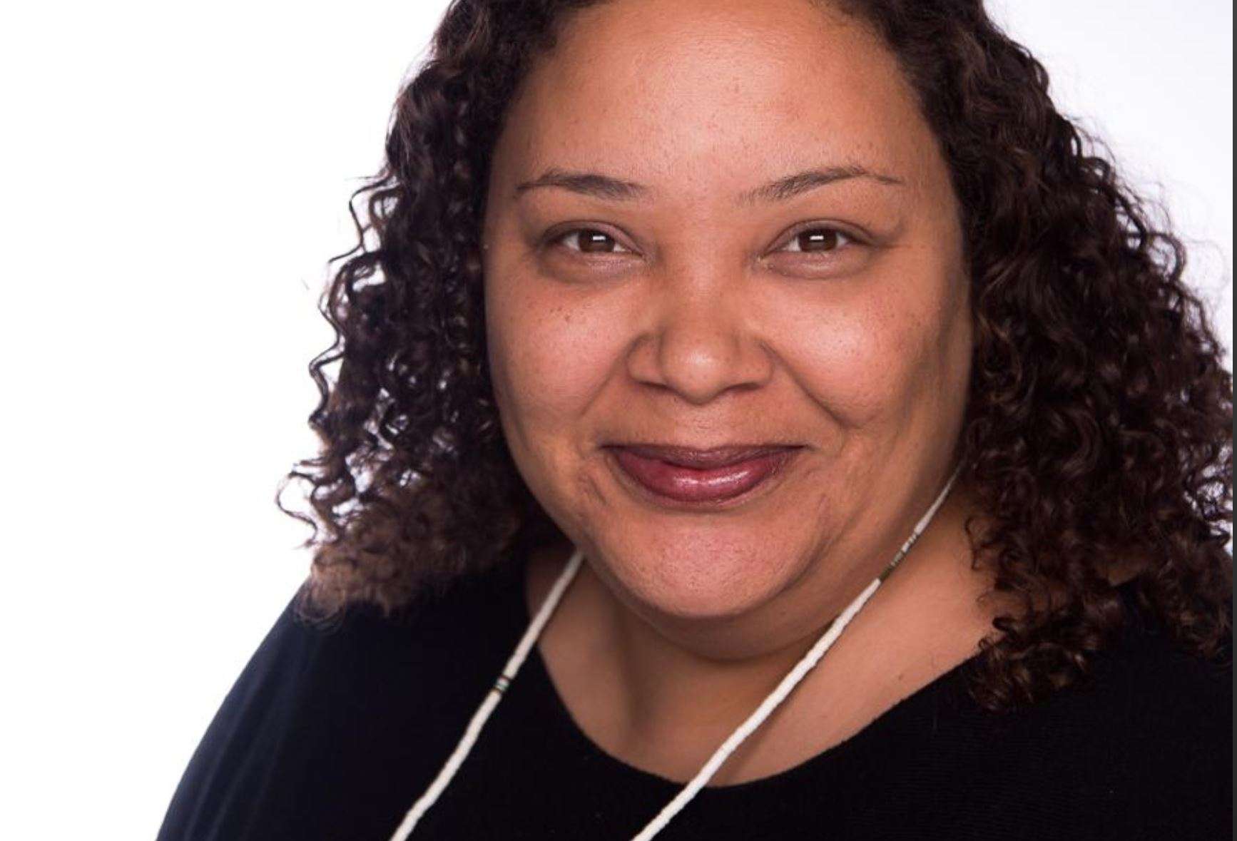 A woman with curly hair smiles warmly, wearing a black shirt and a white-beaded necklace against a plain white background.