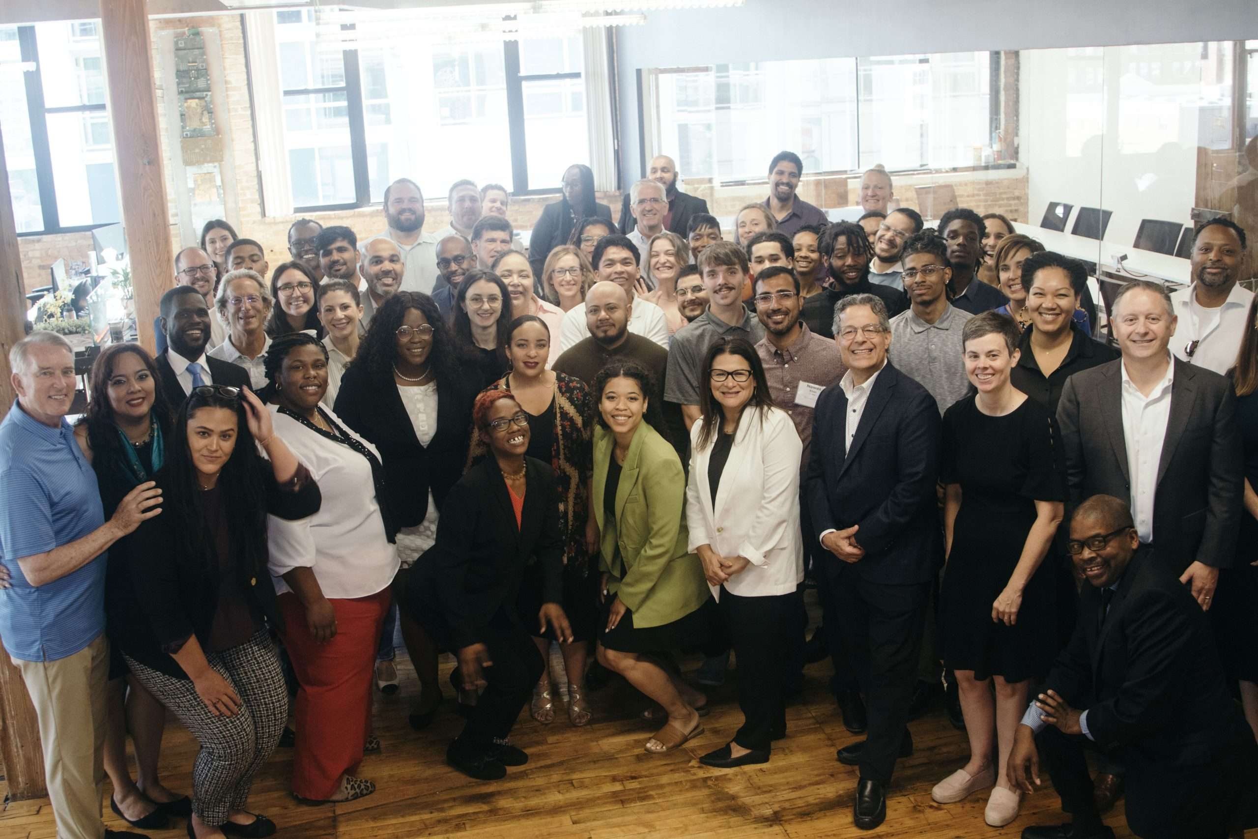 Group of diverse adults standing and posing together, many smiling, in a well-lit office space with large windows and wooden floors in a professional environment.