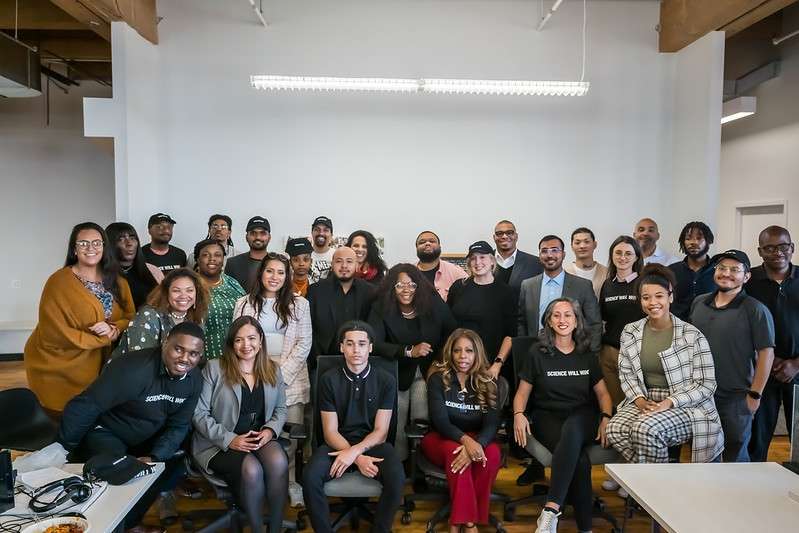 A diverse group of individuals pose smiling in a spacious room with wooden beams. Some are seated while others stand behind wearing casual and business attire; two shirts read "SCIENCE WILL WIN."