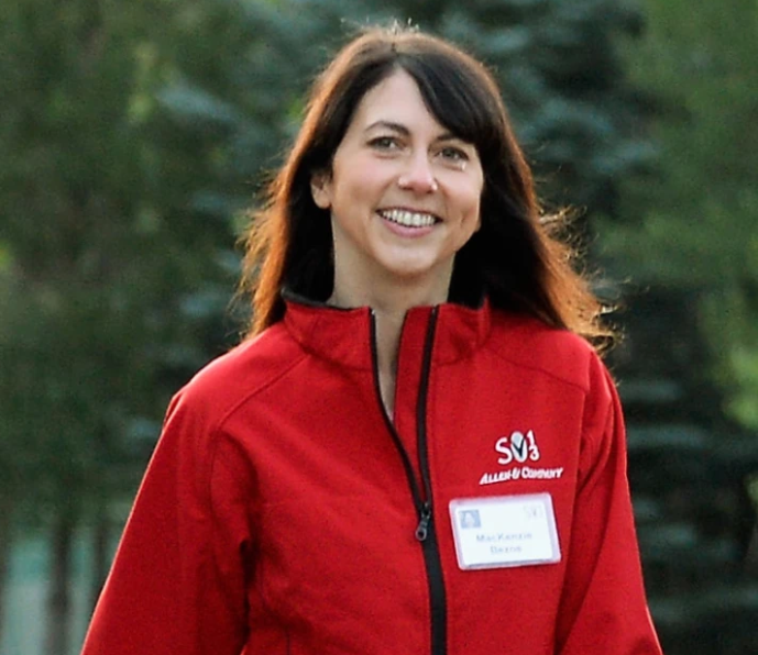 A woman smiling, wearing a red jacket and a name tag reading "MacKenzie Bezos," standing outdoors with green trees in the background.