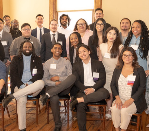 A diverse group of 19 people is posing and smiling indoors, many wearing nametags. Some are seated while others stand behind them, all in a warmly lit room with wooden floors and a window in the background.