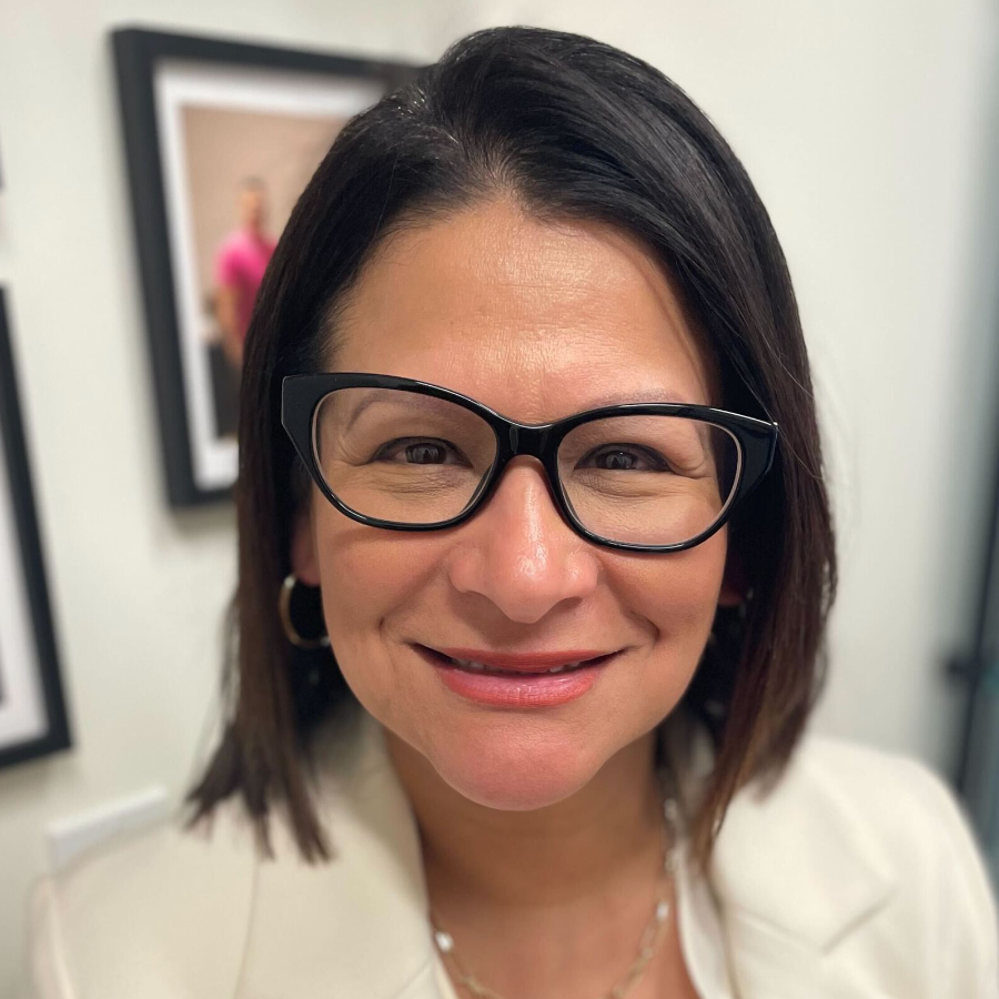 A woman with short dark hair and glasses smiles in an indoor setting, wearing a light-colored blazer, with framed pictures on a white wall in the background.
