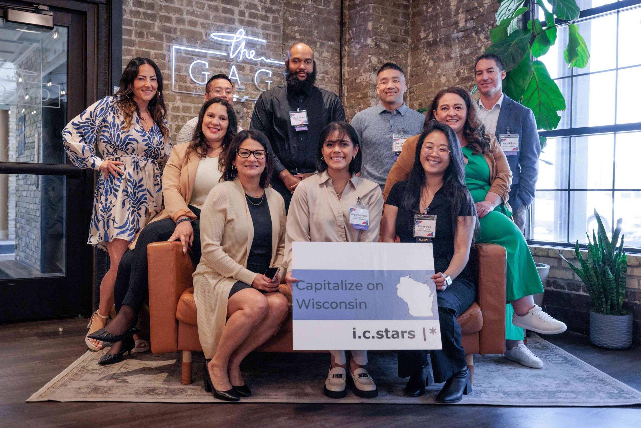 A group of ten people pose indoors, some seated on a couch, holding a sign reading "Capitalize on Wisconsin i.c.stars". Behind them, a brick wall with a neon sign reads "the G.A.C."