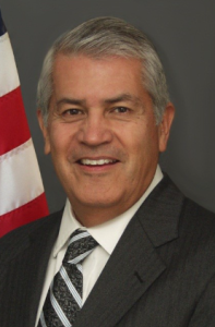 Bill A man with gray hair and a suit smiles in front of an American flag and a plain dark background.