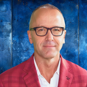 Todd McLees Headshot-3 A man wearing glasses and a red checkered suit jacket stands against a textured blue wall with subtle artistic patterns.