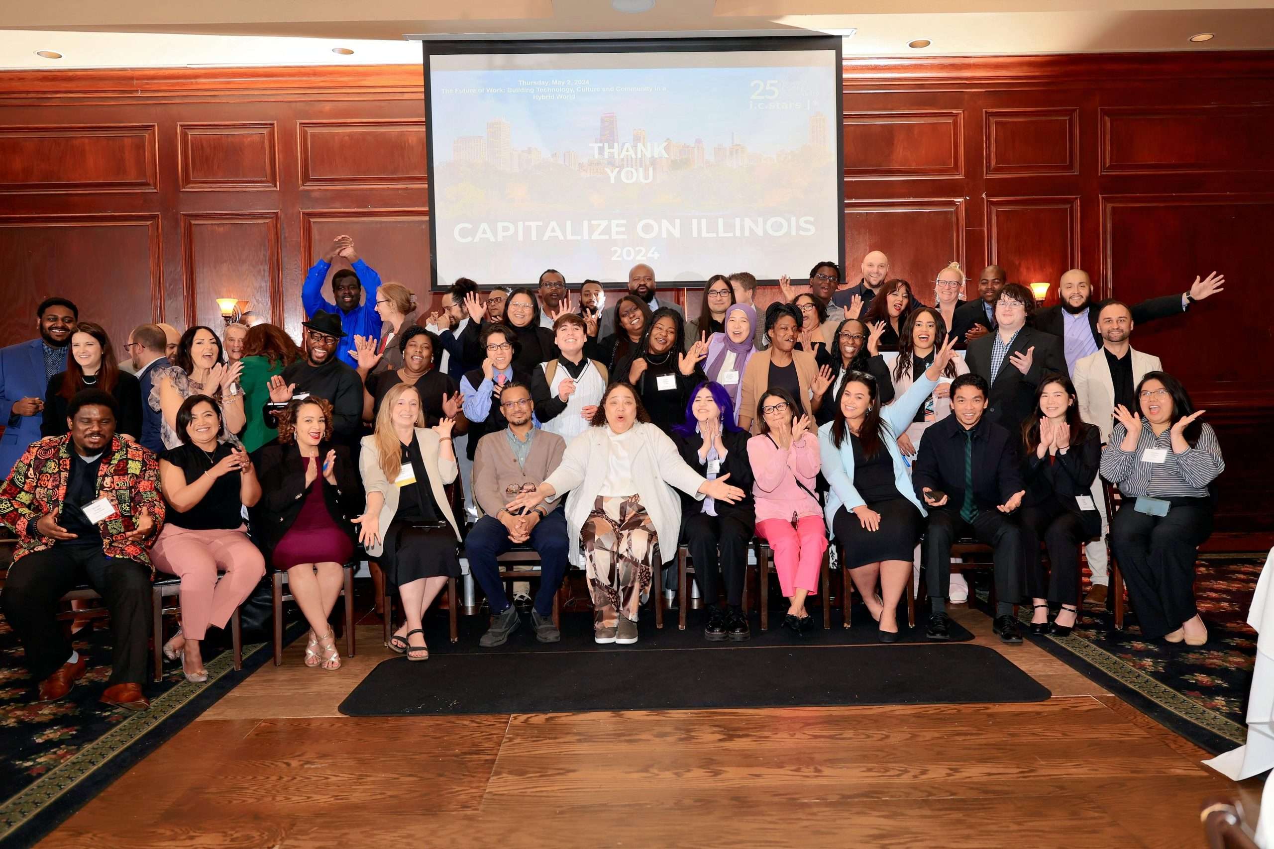 A large group of people enthusiastically posing in front of a wooden wall with a projection screen displaying text, "THANK YOU CAPITALIZE ON ILLINOIS 2024." Everyone is smiling and making joyful gestures.