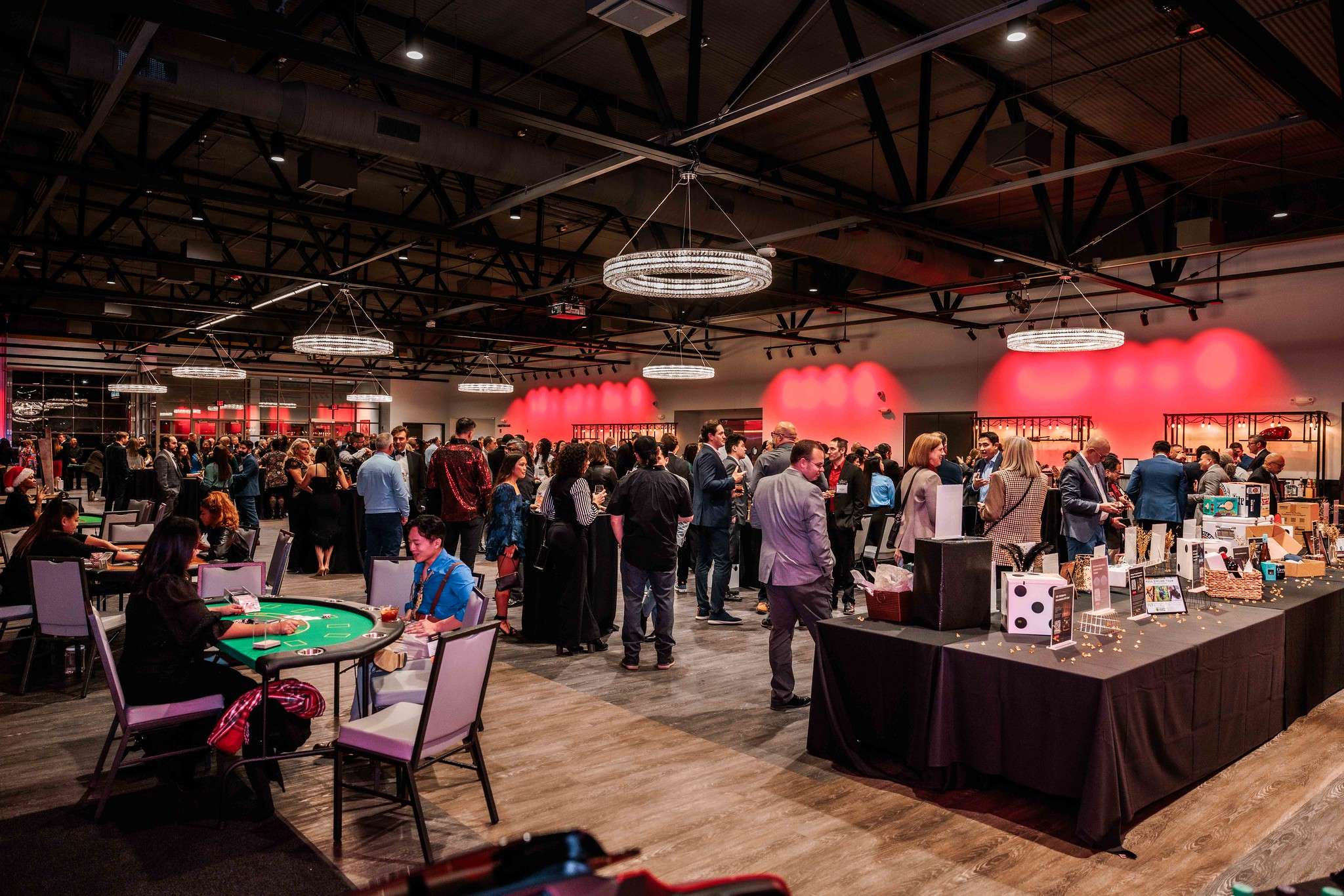 A vibrant scene at Starbash 2024, featuring attendees mingling and engaging in activities like casino games, surrounded by tables with raffle items and illuminated by modern chandeliers in a spacious venue with red accent lighting.