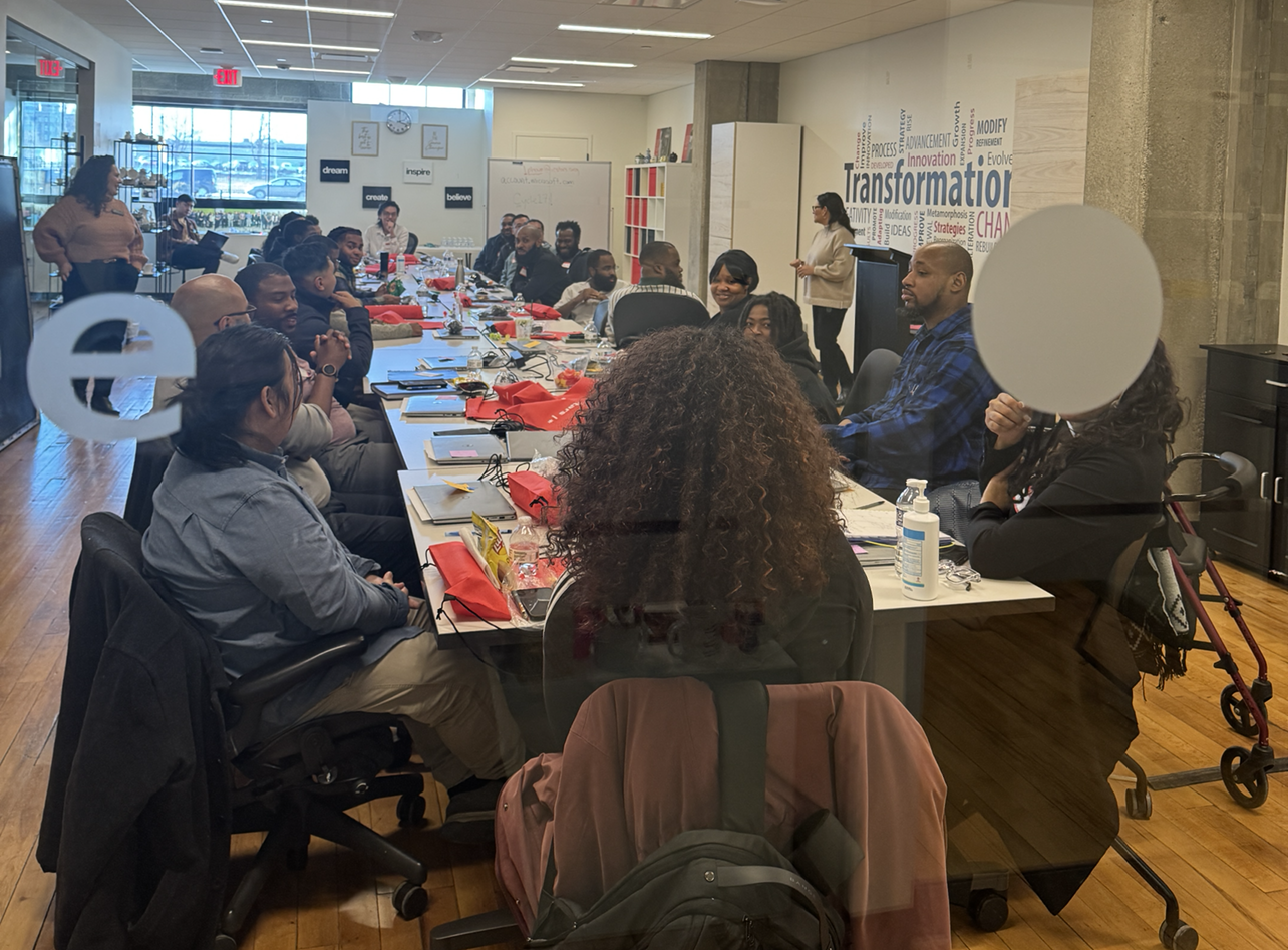 A group of interns seated around a large conference table in a training room. They are engaged in a discussion, with notebooks, folders, and red gift bags on the table. Inspirational words like "Transformation" are displayed on the wall in the background.